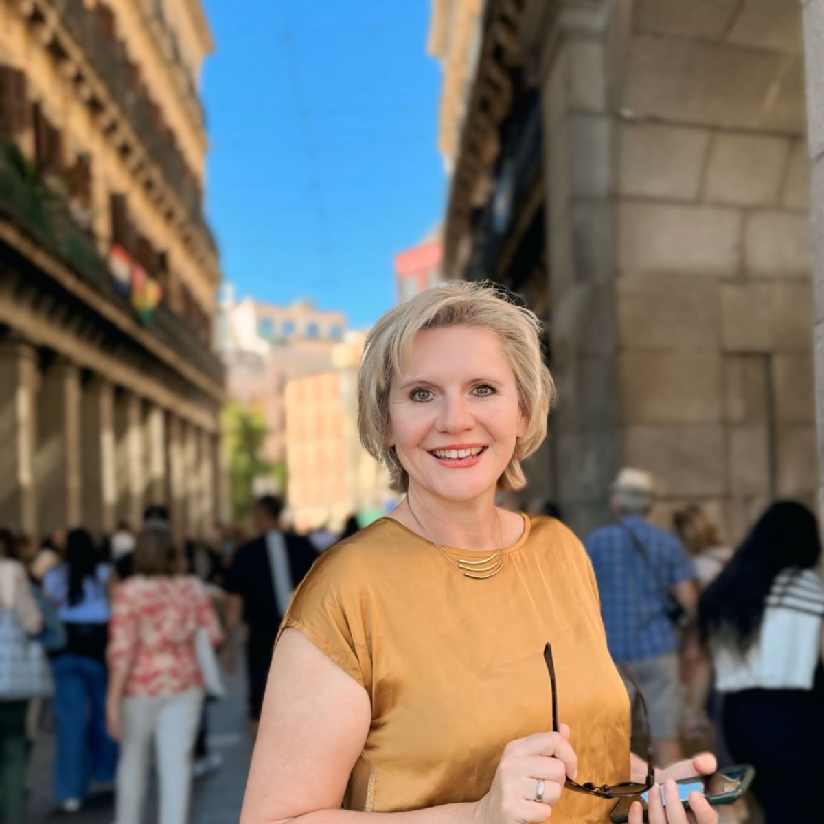Ulli Lang lächelt in die Kamera, sie steht in Madrid am Plaza Major, sie träg ein gelbes Shirt und eine Sonnenbrille in der Hand.
