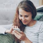 Junge Frau mit langen, braunen Haaren, die lächelnd auf dem Sofa sitzt und chattet.